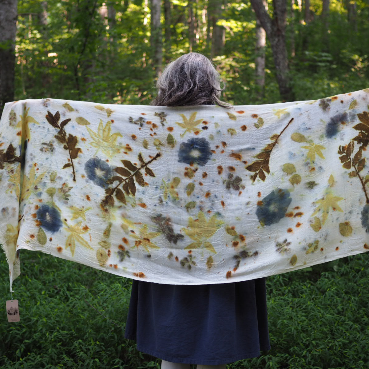 A woman stands in a forest holding open a white woolen shawl ecoprinted with leaves and flowers.