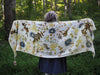 A woman stands in a forest holding open a white woolen shawl ecoprinted with leaves and flowers.