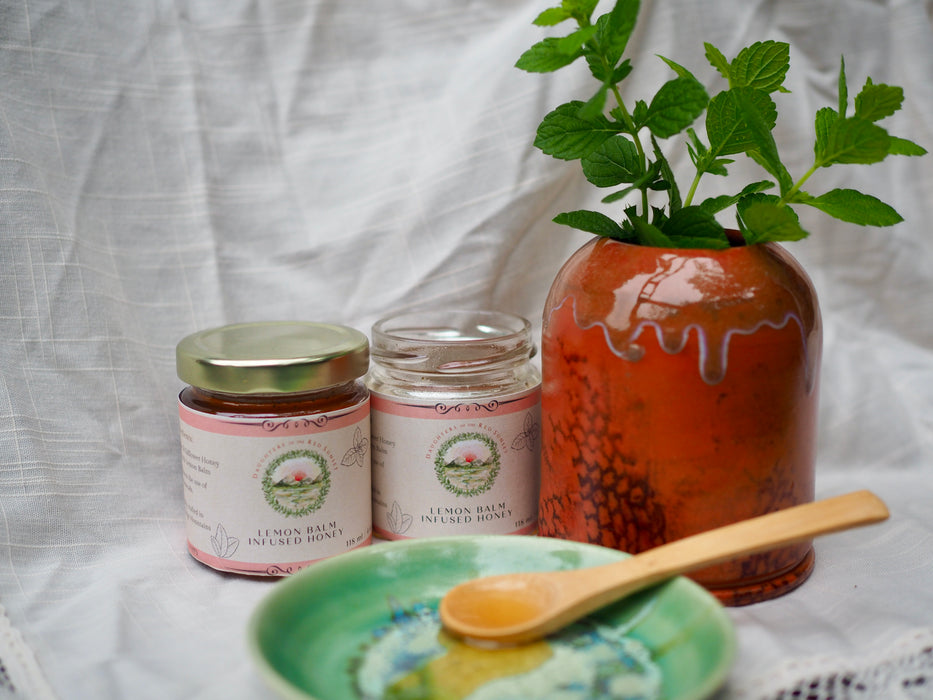 Two jars of honey sit on a lace-edged cloth.  One bottle is open and honey has been spooned onto a small green ceramic plate.  There is Lemon Balm in a terracotta vase next to the honey.