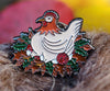 An enamel pin with a cartoon chicken sitting on eggs and red balls of yarn.  Her nest is made of brown and green leaves.