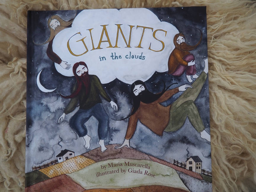 A Giants in the Clouds book sits atop a white sheep fleece.  The book shows a large cloud and four giants dancing on the land under the cloud.