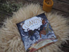 A Giants in the Clouds book sits atop a white sheep fleece, with an orange beeswax candle in the corner. The book shows a large cloud and four giants dancing on the land under the cloud.
