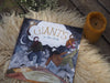 A Giants in the Clouds book sits atop a white sheep fleece, with an orange beeswax candle in the corner.  The book shows a large cloud and four giants dancing on the land under the cloud.
