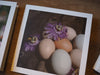 A note card with a photo of six chicken eggs.   On the eggs are two passion flowers with bumble bees flying around them.