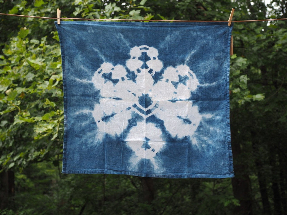 A square blue tea towels hangs on a drying line. The towel has a six petaled design dyed onto it.