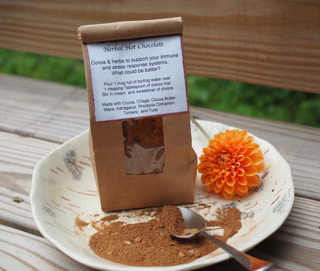 A small brown bag of herbal hot chocolate sits on a white pottery plate with powdered hot chocolate on it.  An orange Dahlia flower sits to the right of the cocoa.